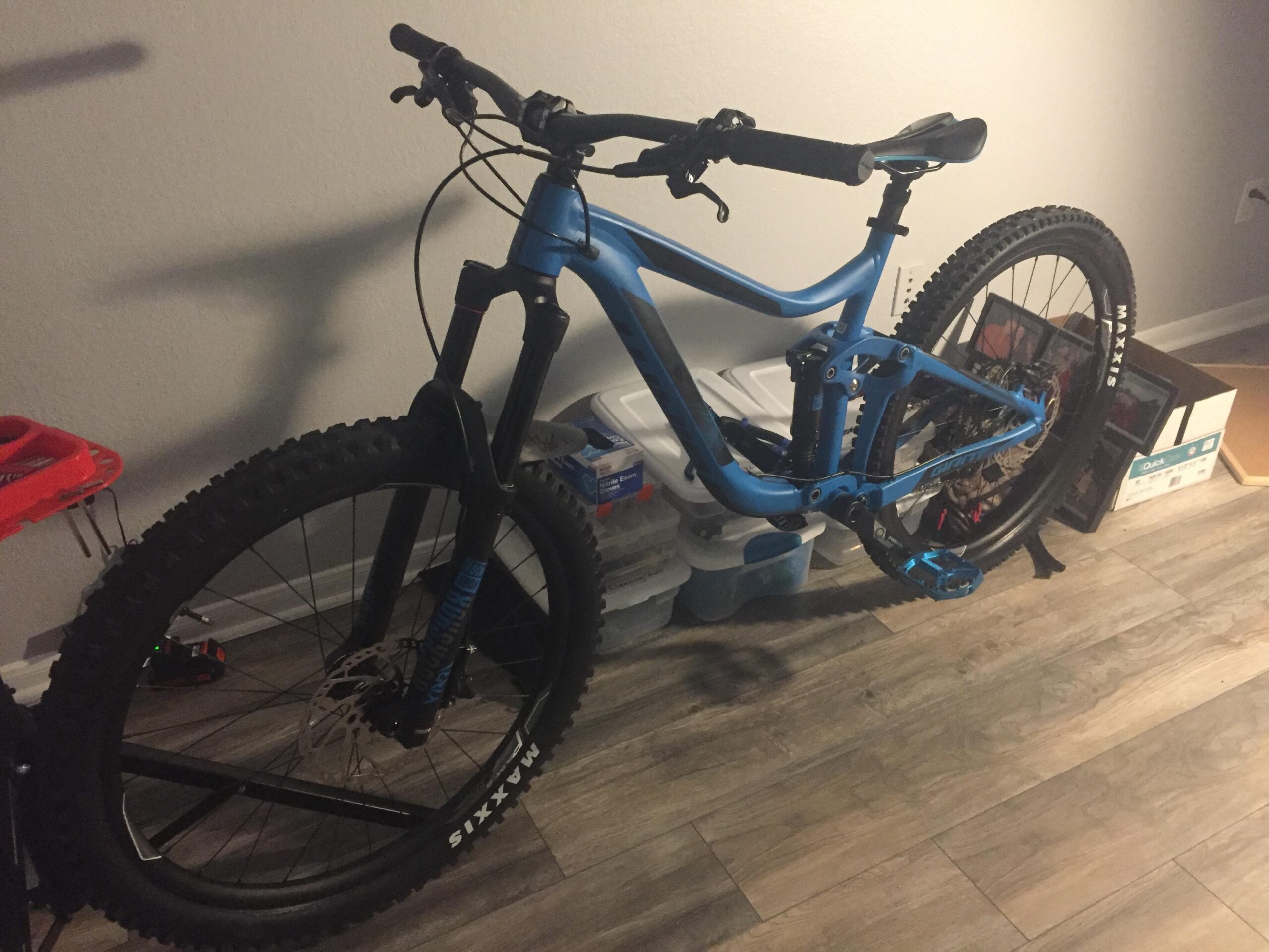 Giant Reign: A blue mountain bike leaning against a wall in a room, with a dark front fork and thick, textured tires. The background features storage containers and a frame, suggesting an indoor environment.