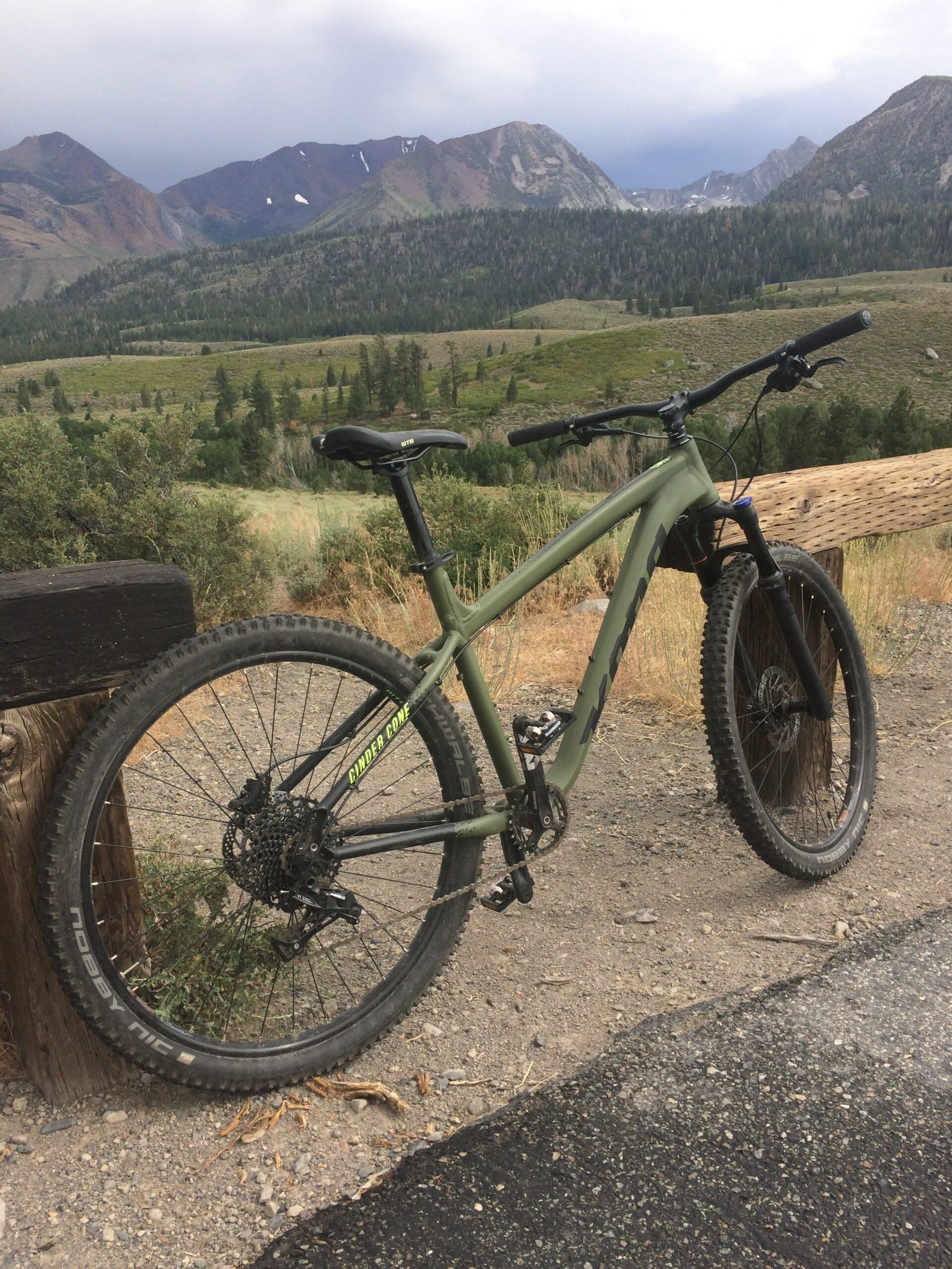 Kona Cinder Cone: A mountain bike parked on a gravel path, with a scenic view of mountains and a lush green valley in the background under a cloudy sky.