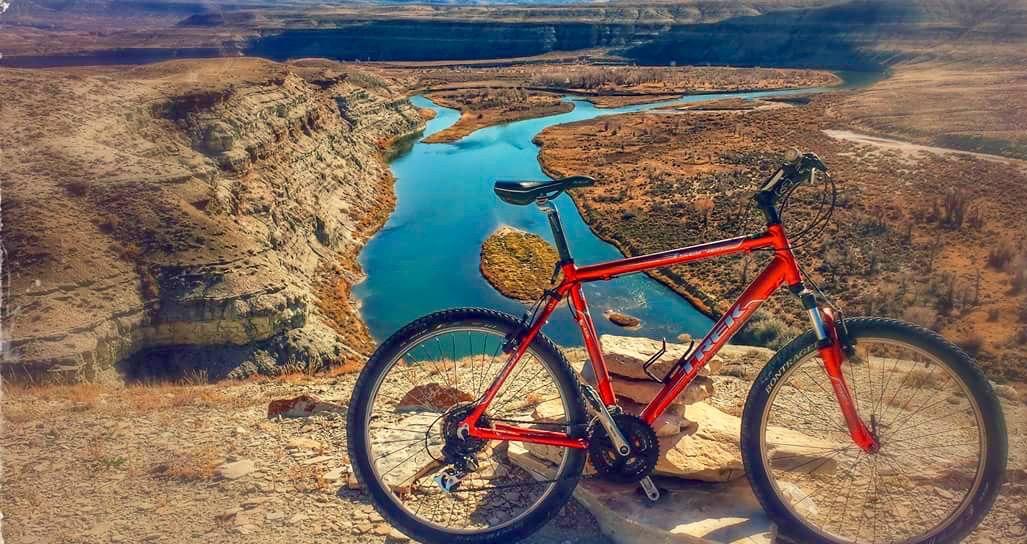 A vibrant red mountain bike leaning against a rocky outcrop, overlooking a winding river surrounded by arid landscapes and cliffs under a clear blue sky. Wilkins Peak Trails mountain bike trail.