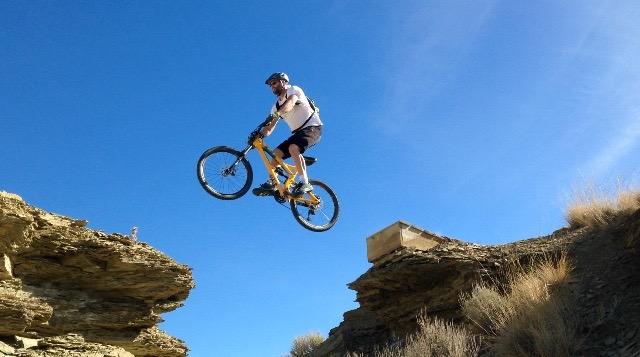 A mountain biker performing a jump over a gap between two rock formations under a clear blue sky. Wilkins Peak Trails mountain bike trail.
