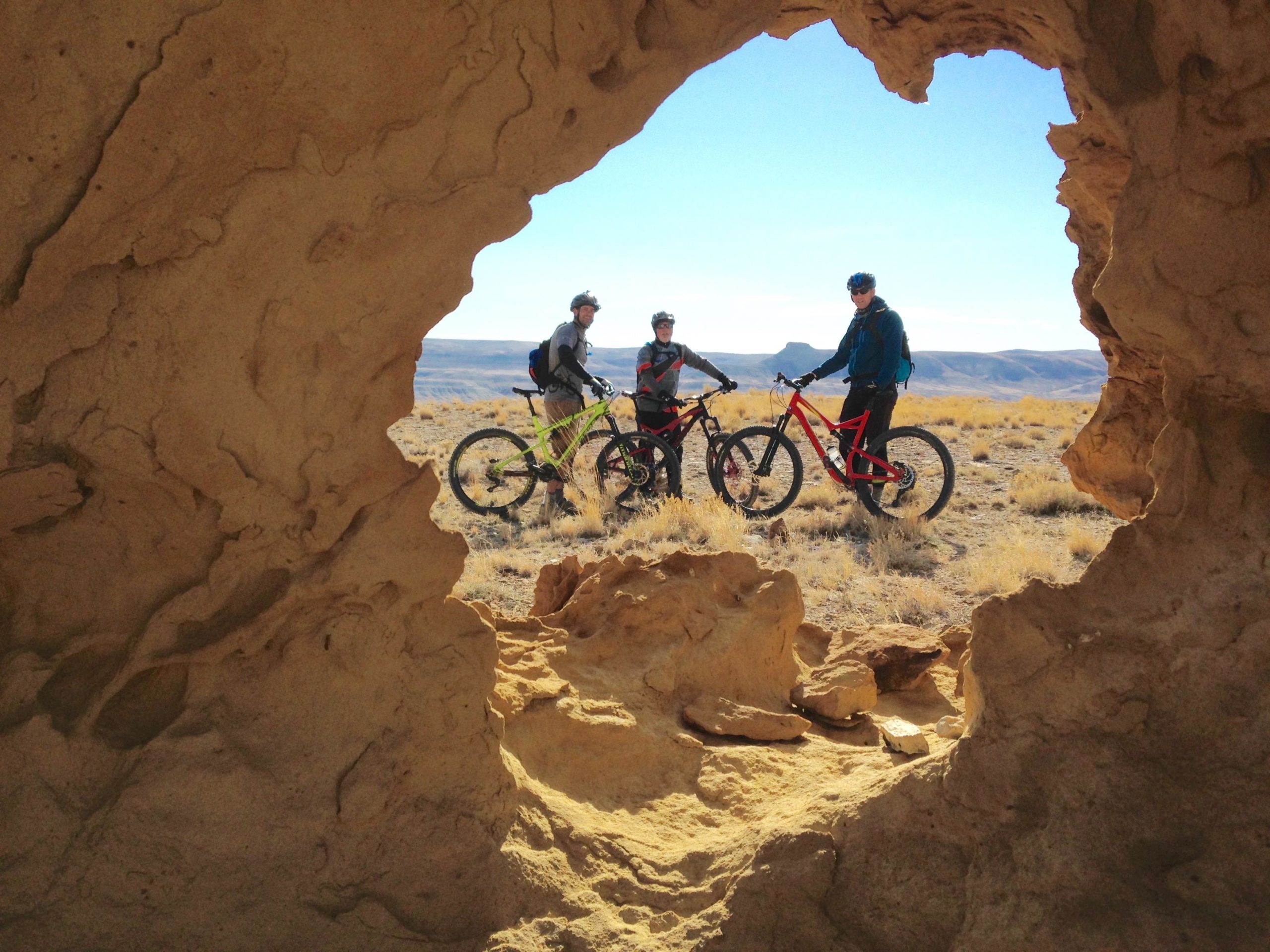 Three mountain bikers standing with their bikes in a dry, rocky landscape, viewed through a natural rock formation. The area is sparsely vegetated, with hills visible in the background under a clear blue sky. Wilkins Peak Trails mountain bike trail.