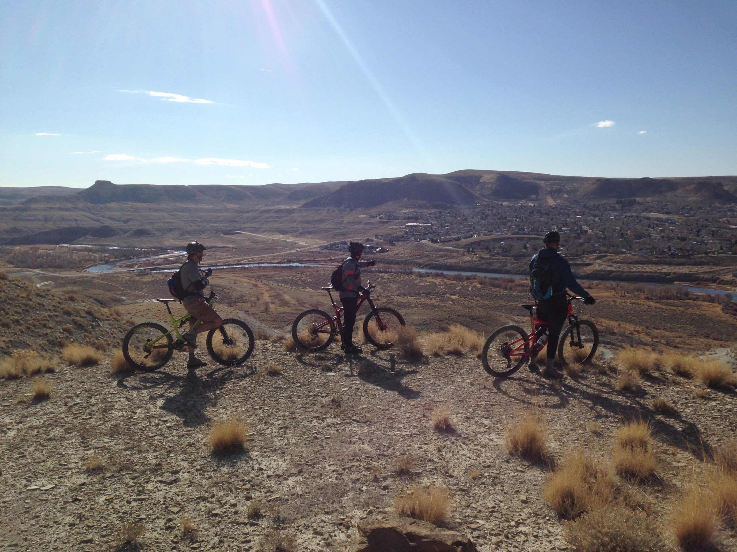 Three mountain bikers stand on a rocky overlook, gazing at a valley below. The landscape features rolling hills, a winding river, and a small town in the distance under a clear blue sky with a few clouds. The sun casts a warm glow on the scene. Wilkins Peak Trails mountain bike trail.