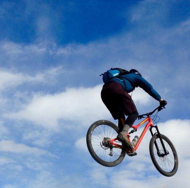 A cyclist performing a jump on a mountain bike against a backdrop of blue sky with scattered clouds. The rider is wearing a blue shirt and shorts, and is in mid-air with the bike elevated off the ground. Wilkins Peak Trails mountain bike trail.