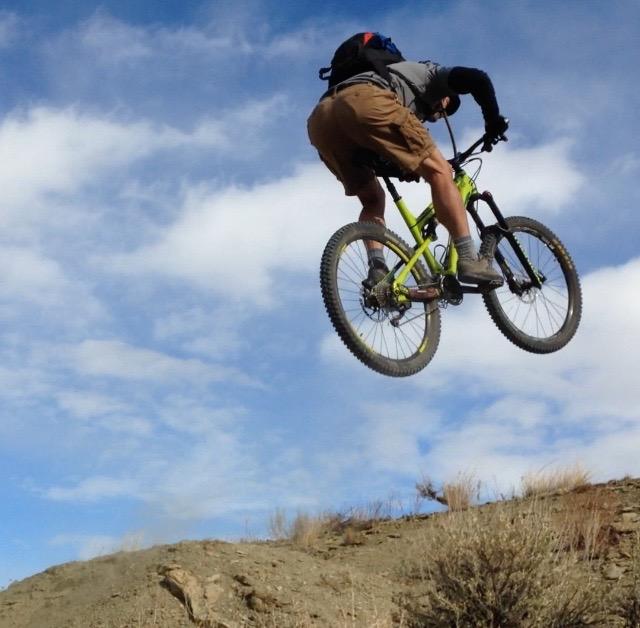 A person in shorts performs a jump on a green mountain bike against a backdrop of blue sky with scattered clouds. The rider is airborne, with their body positioned upward, capturing the thrill of extreme biking. The terrain is rocky and dry, suggesting an outdoor biking trail. Wilkins Peak Trails mountain bike trail.