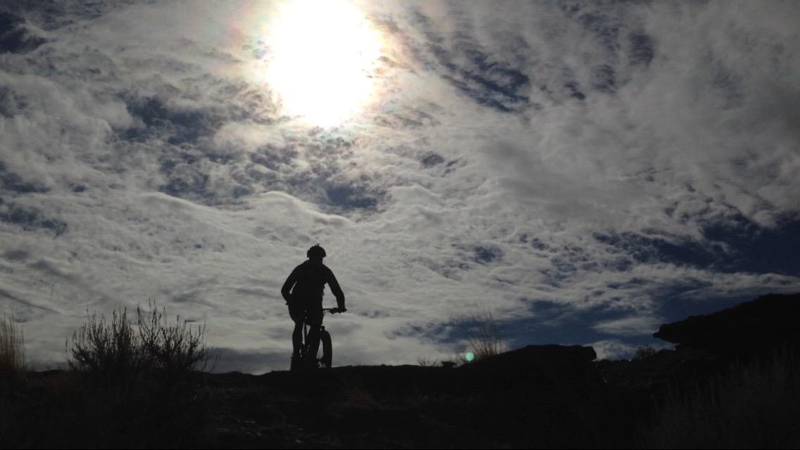 A silhouette of a mountain biker against a dramatic sky filled with clouds, with the sun shining brightly above. The scene captures a sense of adventure and the beauty of an outdoor landscape. Wilkins Peak Trails mountain bike trail.