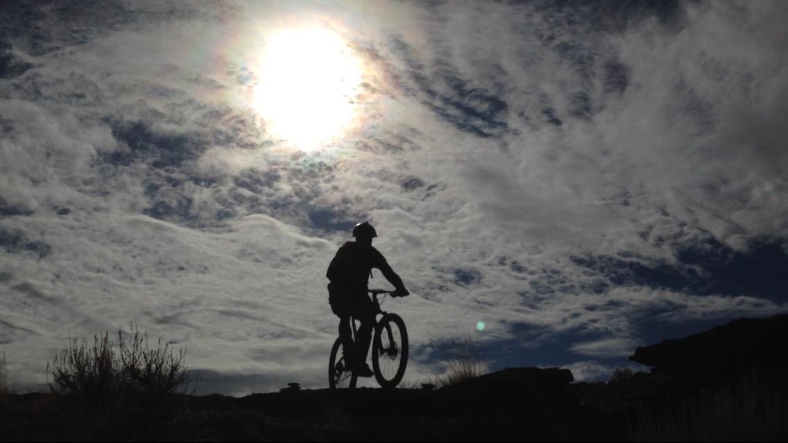 A silhouette of a cyclist riding on rocky terrain against a backdrop of dramatic clouds and a bright sun. The scene captures the essence of outdoor adventure and vibrant nature. Wilkins Peak Trails mountain bike trail.