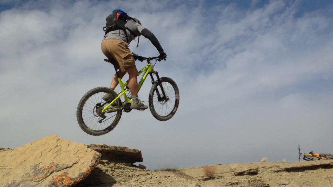 A person on a mountain bike is performing a jump over a rocky ledge against a backdrop of a blue sky with clouds. The cyclist is wearing a helmet and a backpack, displaying a dynamic pose mid-air. Wilkins Peak Trails mountain bike trail.