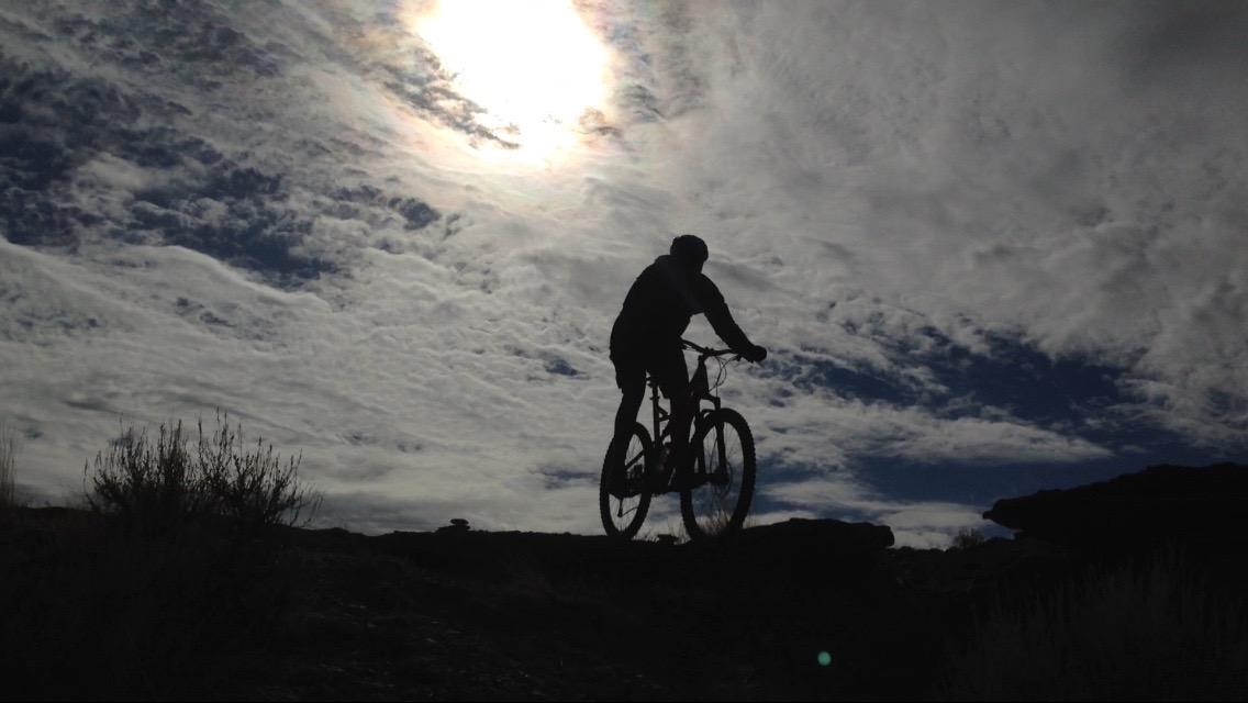Silhouette of a mountain biker riding on a rocky trail against a backdrop of dramatic clouds and a bright sun in the sky. Wilkins Peak Trails mountain bike trail.