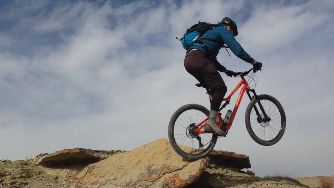 A mountain biker performing a jump off a rocky outcrop against a backdrop of cloudy blue sky. The cyclist is wearing a helmet and a backpack, showcasing action and adventure in outdoor sports. Wilkins Peak Trails mountain bike trail.