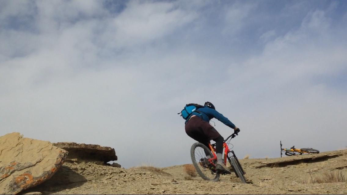 A mountain biker ascending a rocky trail under a partially cloudy sky, with another bike lying on the ground nearby. Wilkins Peak Trails mountain bike trail.