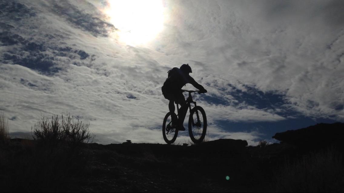 A silhouette of a cyclist riding a mountain bike on rocky terrain, with the sun shining brightly in the background and dramatic clouds in the sky. Wilkins Peak Trails mountain bike trail.