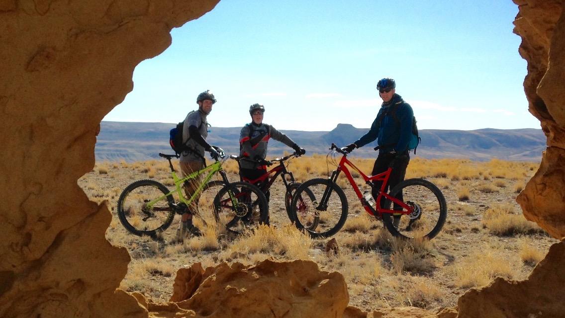 Three mountain bikers stand beside their bikes in a desert landscape, framed by a rock formation. The riders, dressed in outdoor gear, are smiling and posing for the photo, with the blue sky and distant hills visible in the background. Wilkins Peak Trails mountain bike trail.