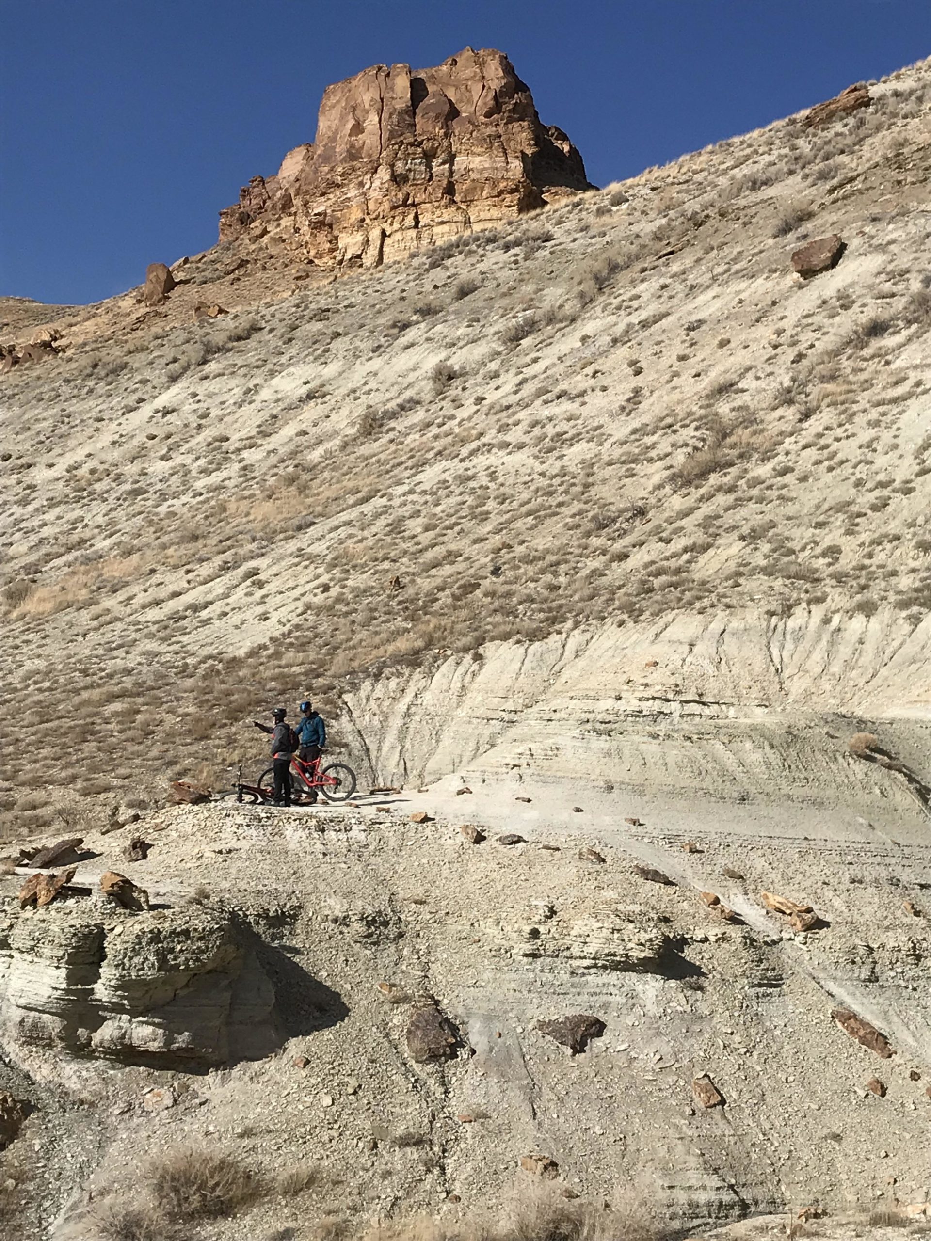 Two mountain bikers pause on a rocky terrain in a semi-arid landscape, with a prominent rock formation towering in the background under a clear blue sky. The scene captures a moment of exploration and adventure in nature. Wilkins Peak Trails mountain bike trail.