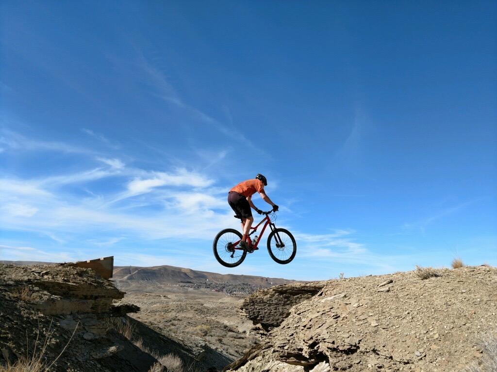 A mountain biker in an orange shirt and helmet jumps off a rocky ledge against a clear blue sky, showcasing an adventurous moment in a rugged outdoor setting. Wilkins Peak Trails mountain bike trail.