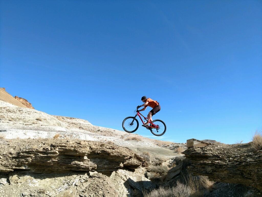 A mountain biker in an orange shirt and black shorts jumps off a rocky ledge against a clear blue sky, showcasing an adventurous moment in a rugged outdoor setting. Wilkins Peak Trails mountain bike trail.