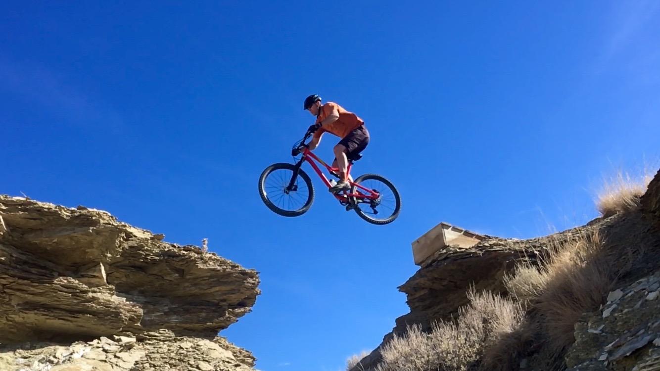 A mountain biker jumps over a rocky gap, soaring in mid-air against a clear blue sky. The rider is wearing an orange shirt and a helmet, showcasing a dynamic moment of extreme sports. Wilkins Peak Trails mountain bike trail.