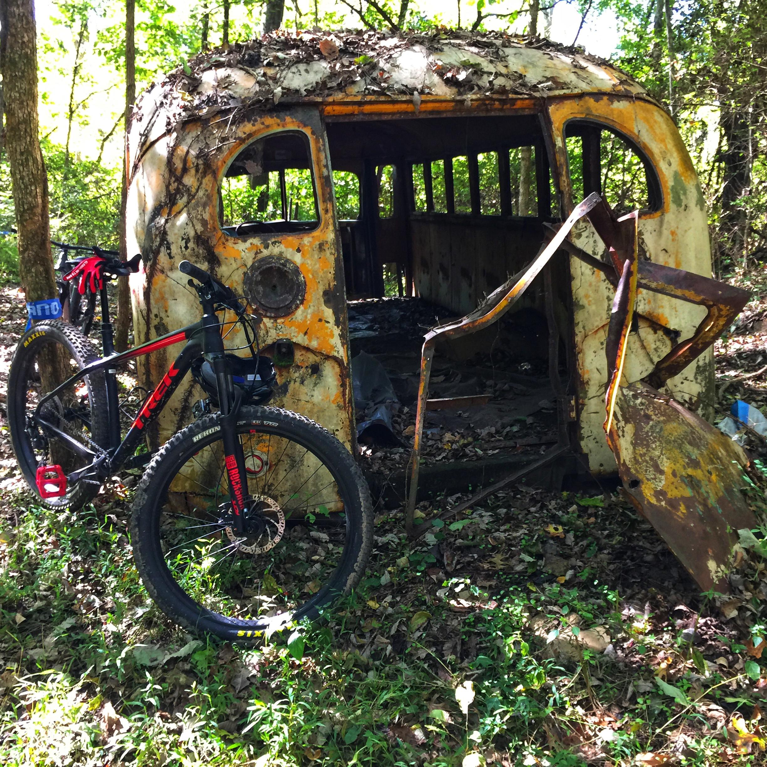 Trek Roscoe 7: A rusty, abandoned vehicle covered in leaves and surrounded by greenery, with a mountain bike positioned beside it. The vehicle's interior is visible, showcasing decayed wood and metal elements. Sunlight filters through the trees, creating a serene yet slightly eerie atmosphere.