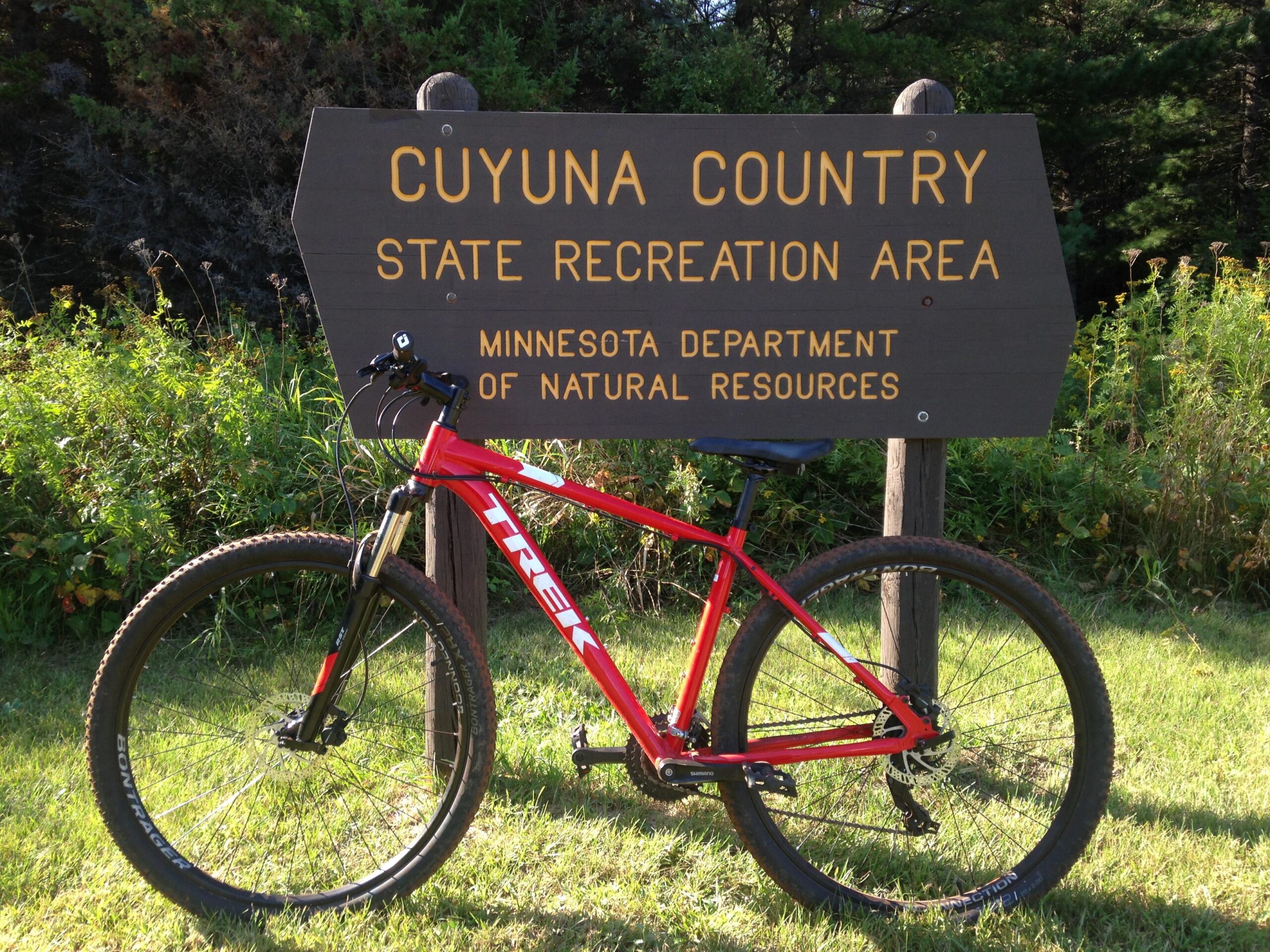 Trek Marlin 5: A red Trek mountain bike leaning against a wooden sign that reads "Cuyuna Country State Recreation Area" with "Minnesota Department of Natural Resources" below it. The background features green grass and trees, suggesting a natural outdoor setting.