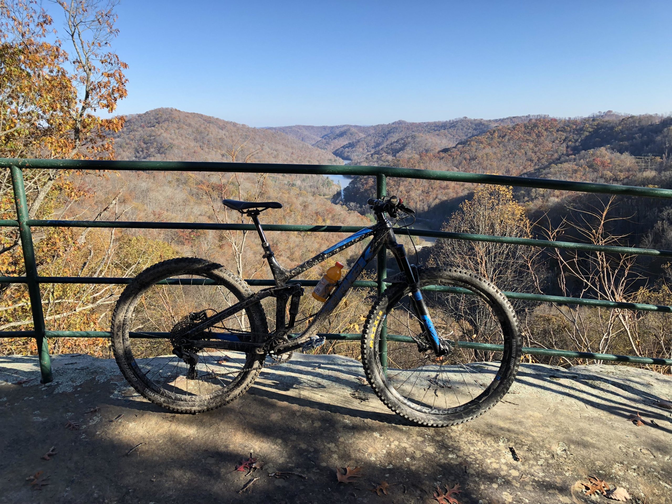 A mountain bike parked at a viewpoint, overlooking a scenic landscape of hills and a river. The surrounding trees display autumn foliage, and the sky is clear blue. A green railing frames the scene, enhancing the sense of depth in the natural setting. Sugarcamp Mountain mountain bike trail.