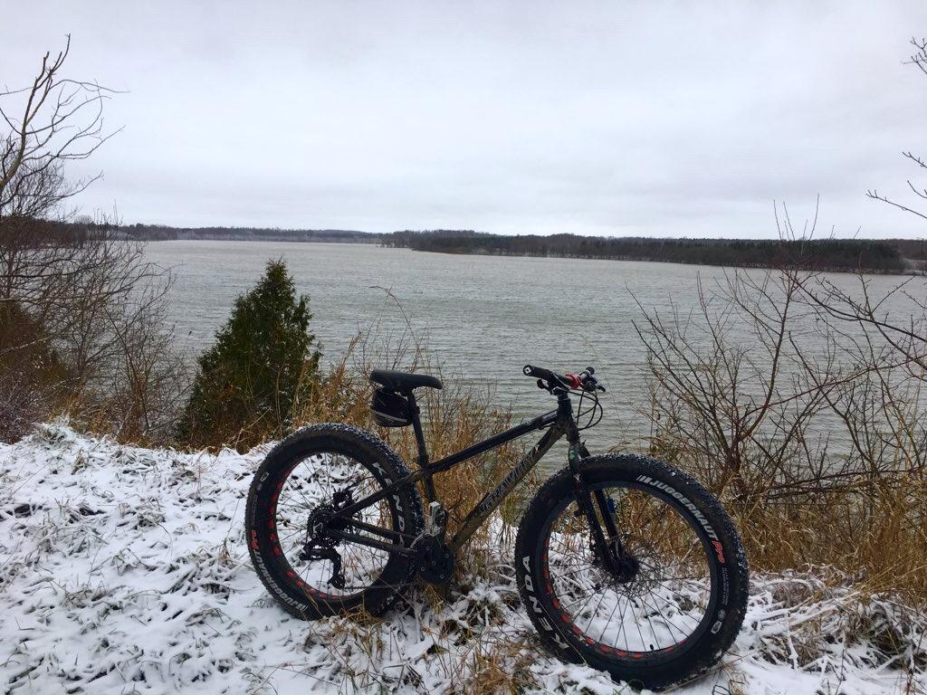 A fat tire bicycle is parked near a snowy bank beside a lake, surrounded by sparse trees and a cloudy sky. The calm water reflects the overcast weather, creating a serene winter scene. Fanshawe Lake mountain bike trail.