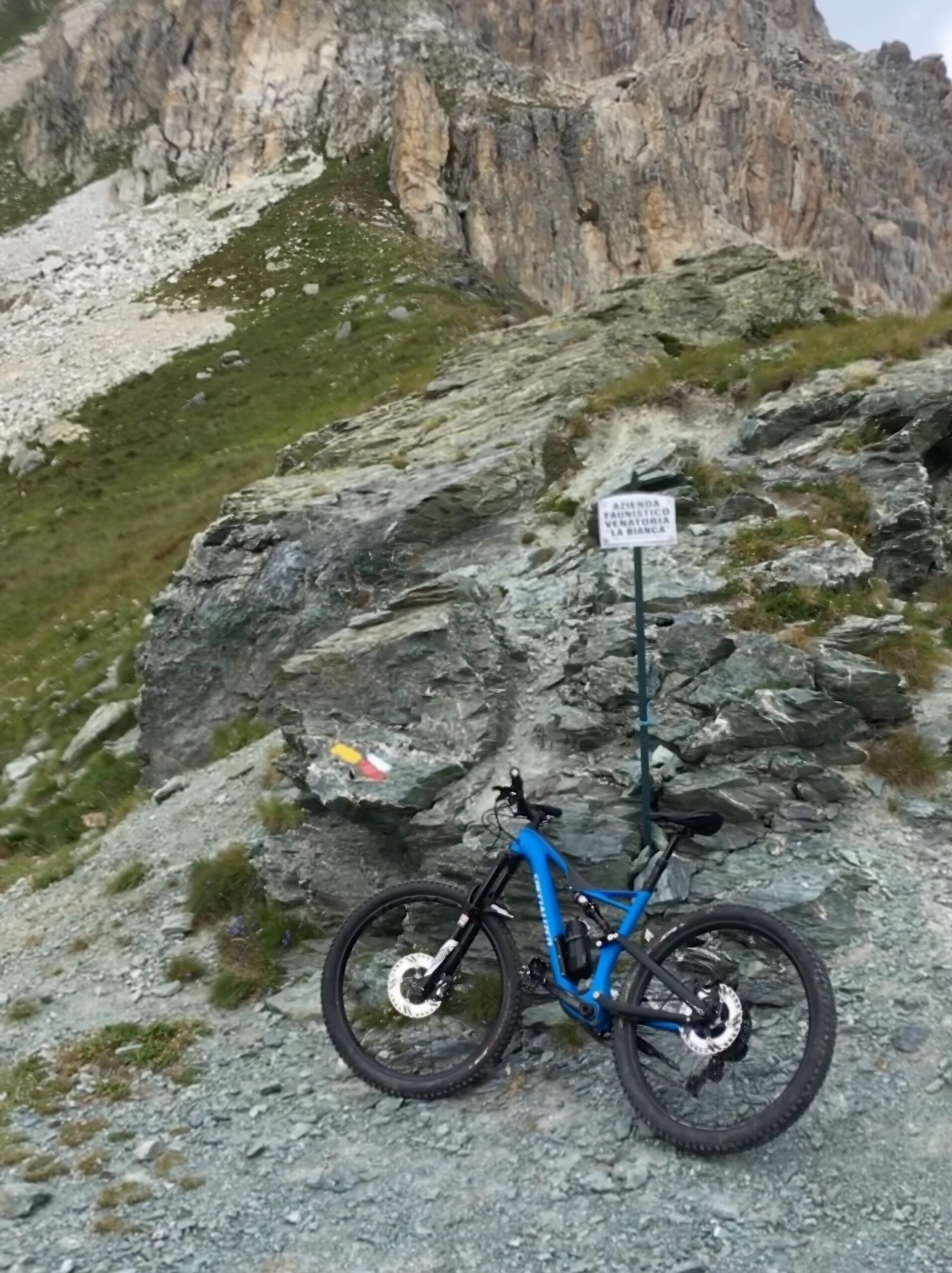 Specialized Stumpjumper FSR comp carbon 650B: A blue mountain bike is leaned against a rocky outcrop in a mountainous area. In the background, there are steep cliffs and grassy slopes. A sign is posted nearby, and colorful trail markers can be seen on the rocks.