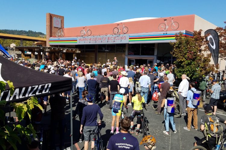 A large crowd gathers in front of the Marin Museum of Bicycling on a sunny day. People of various ages, some wearing cycling gear and helmets, stand in a parking area while others sit on bikes. A couple of individuals are on a stage, addressing the audience. Banners and colorful decorations adorn the museum entrance, and nearby tents indicate it may be a cycling event or festival.