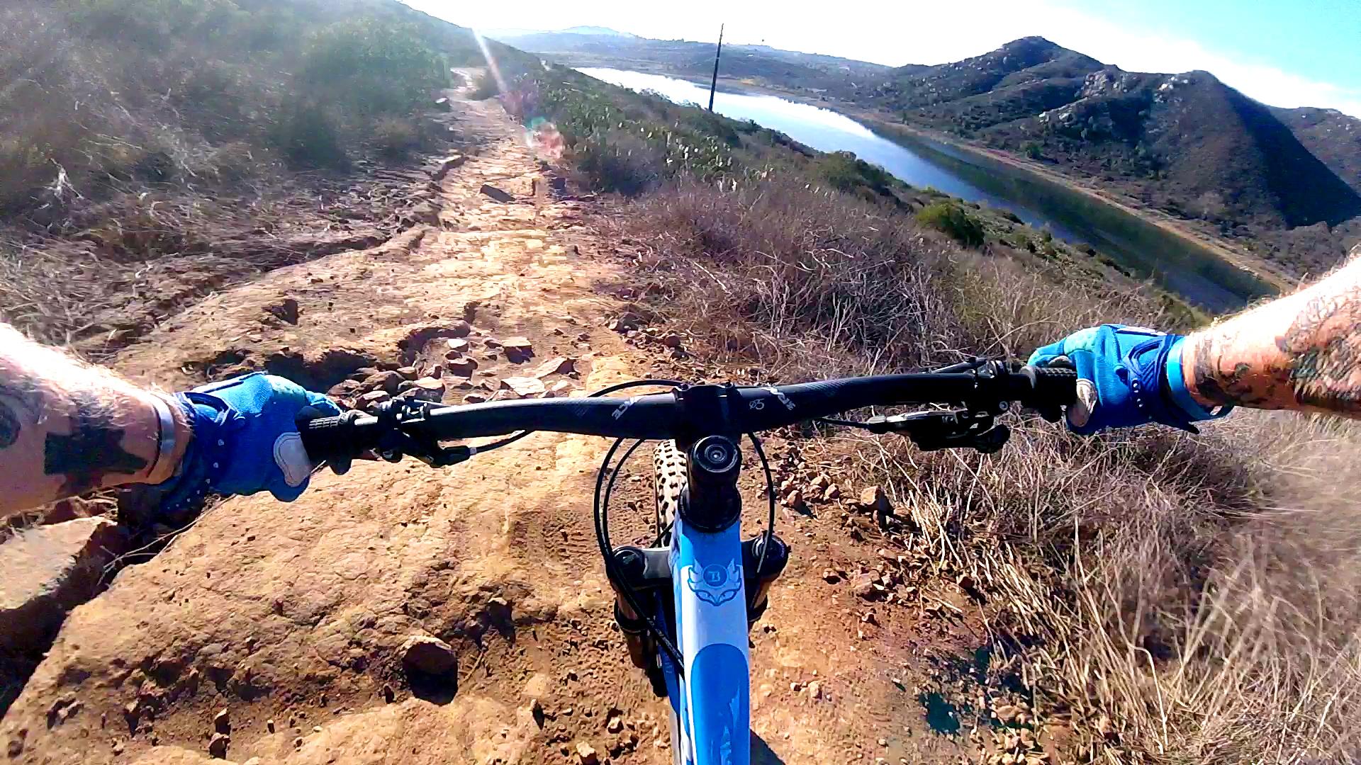 A mountain biker's perspective on a rugged dirt trail with rocky patches, overlooking a serene body of water surrounded by hills. The handlebars of the bike are in focus, and the scene is bright with sunlight filtering through the landscape. Lake Hodges mountain bike trail.