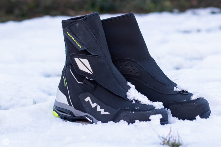 Black winter boots partially covered in snow, featuring sleek design elements and branded with the "Northwave" logo. The boots have a high ankle design and a rugged sole, suitable for cold weather conditions.
