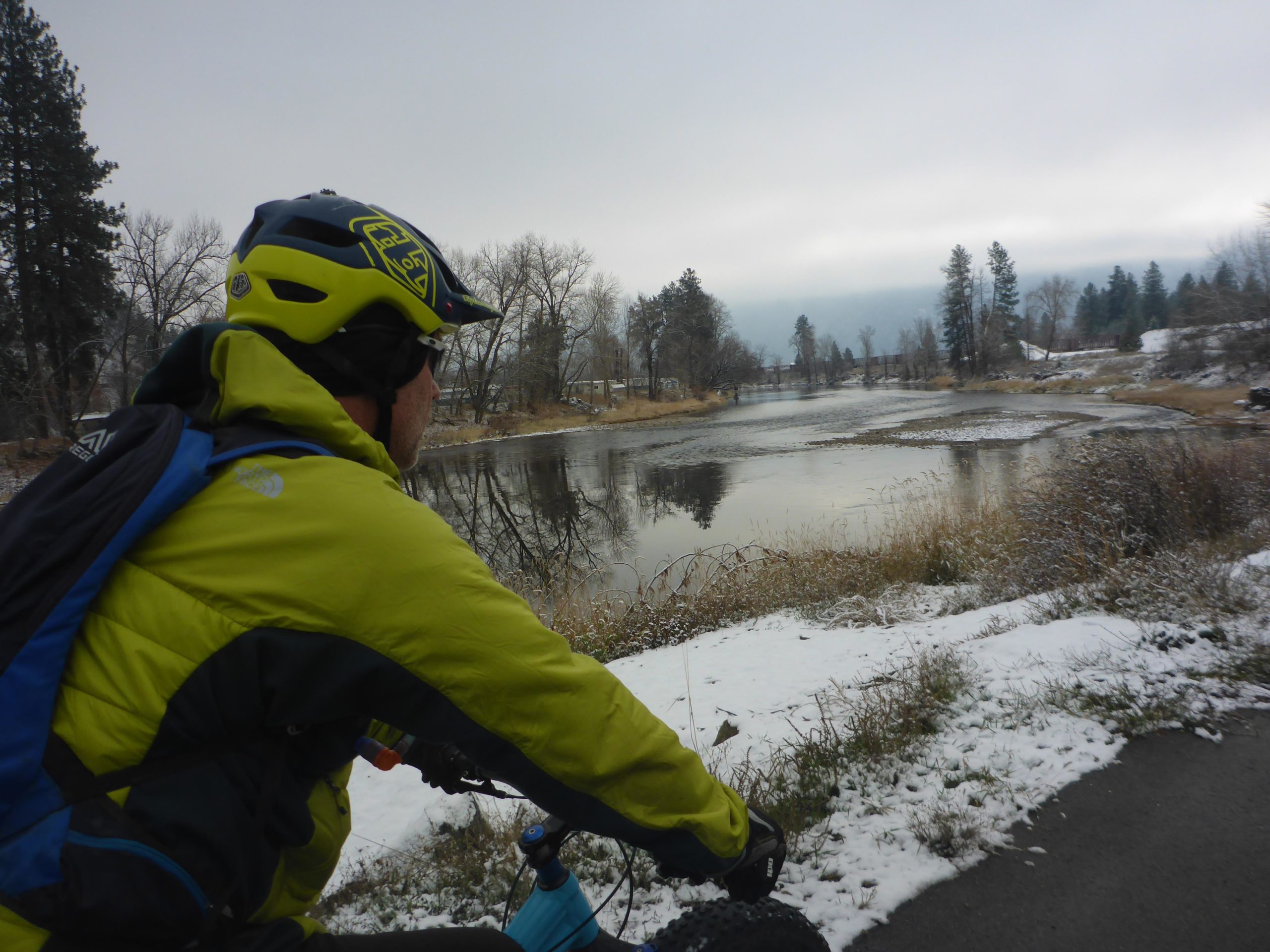 A person wearing a bright yellow jacket and helmet sits on a bicycle, looking out over a calm river surrounded by bare trees and patches of snow. The sky is overcast, creating a serene, wintry landscape. Trans Canada Trail - Grand Forks to Christine Lake mountain bike trail.