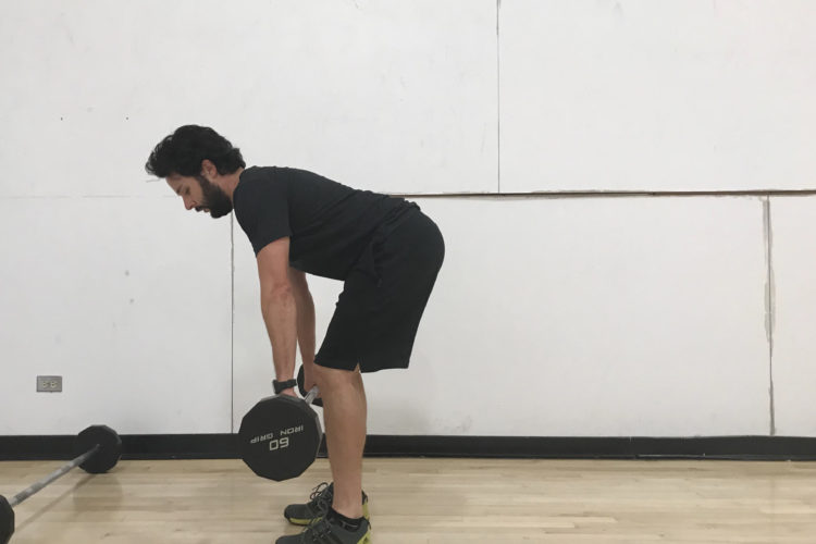 A man in a black shirt and shorts performs a dumbbell deadlift in a gym setting, focusing on his form. He is bent at the hips with a slight forward lean, holding a dumbbell in one hand while a barbell and another dumbbell rest on the floor nearby. The background features a plain white wall and a wooden gym floor.