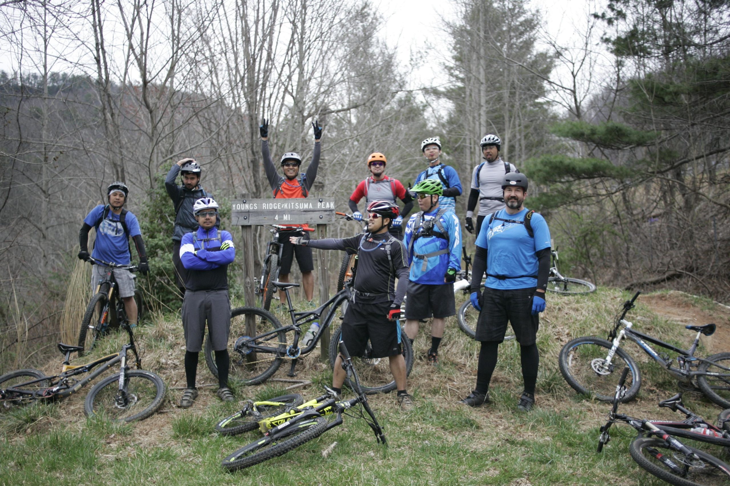 A group of ten mountain bikers posing together at a trailhead sign that reads "Young's Ridge/Kitsuma Peak 4 Mi." The bikers are wearing helmets and cycling gear, with some sitting on their bikes and others standing proudly, displaying various facial expressions ranging from excitement to enthusiasm. The background features trees and a natural landscape, indicating a wooded outdoor setting. Kitsuma mountain bike trail.