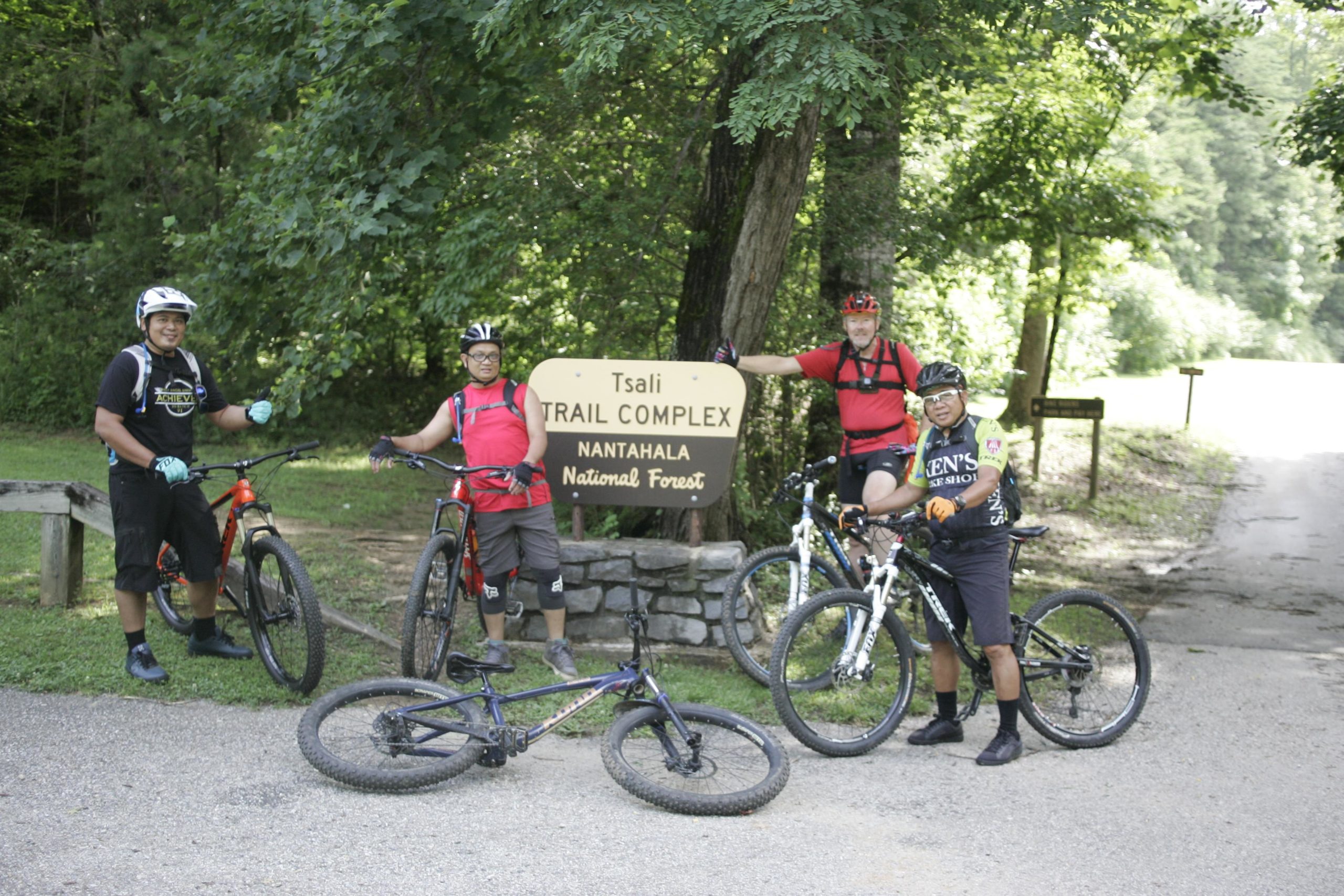Four mountain bikers posing in front of the Tsali Trail Complex sign in Nantahala National Forest, surrounded by greenery. Two bikes are standing upright, while one is lying on the ground. Tsali Left Loop mountain bike trail.