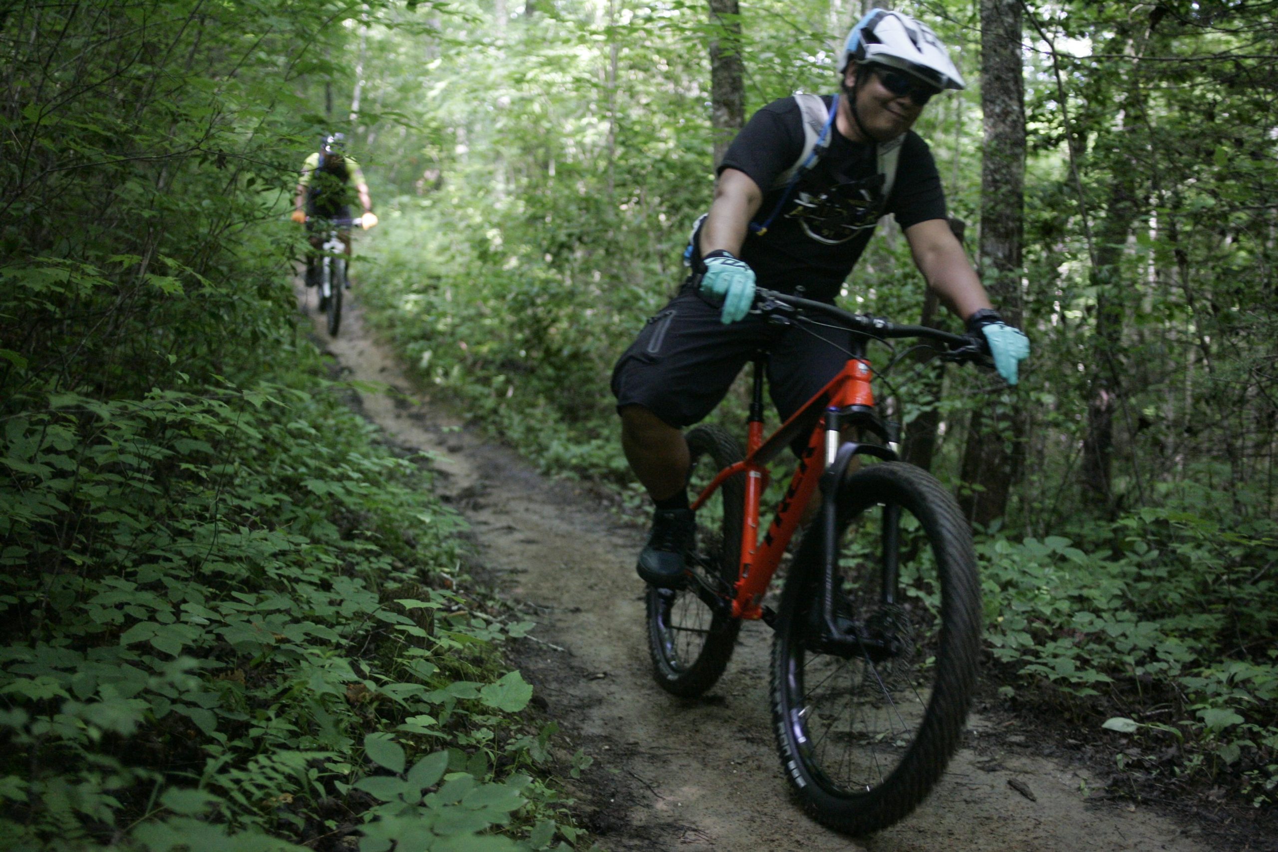 Two mountain bikers riding on a narrow, dirt trail surrounded by lush green vegetation. One rider, wearing a black shirt, shorts, and a helmet, is smiling as he pedals on an orange bike, while the other rider is visible in the background. Tsali Left Loop mountain bike trail.