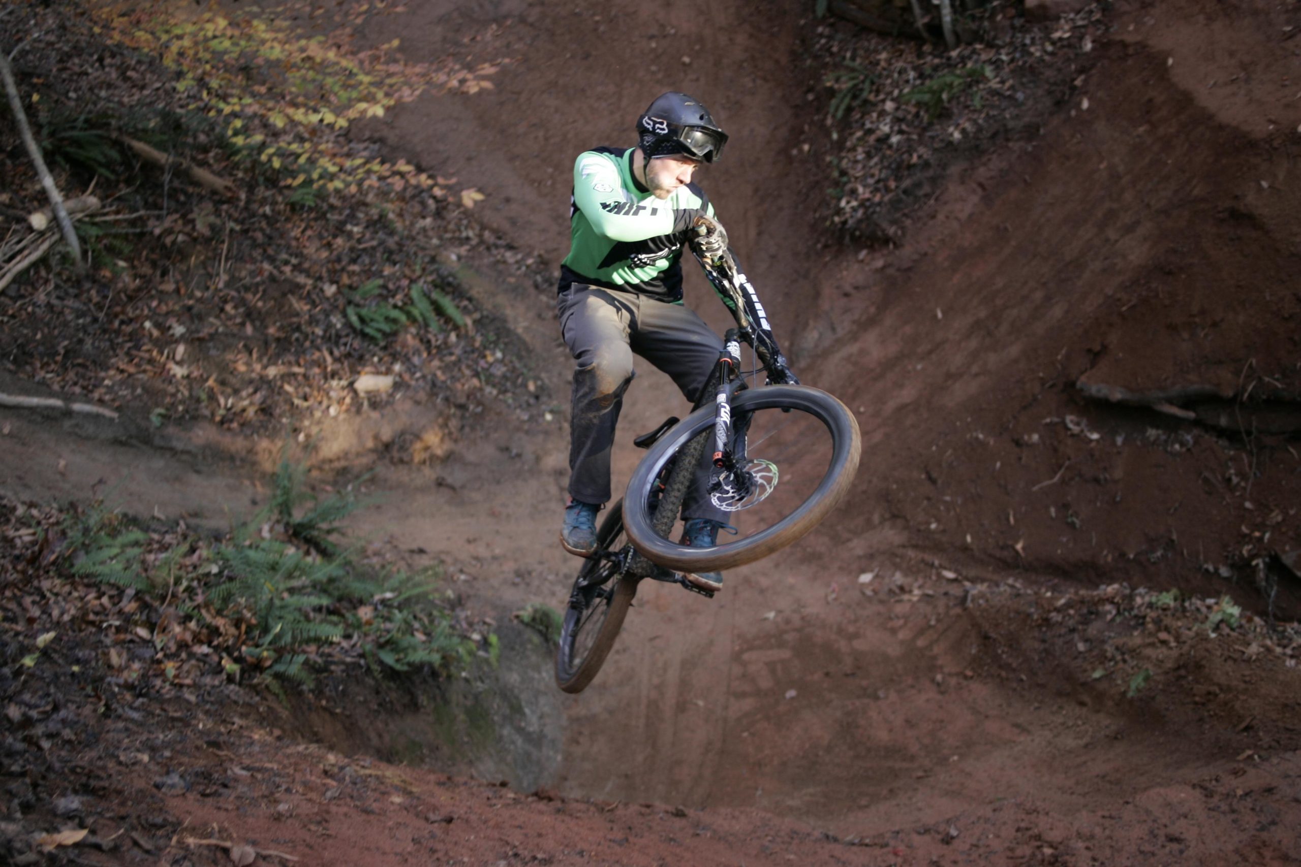 A mountain biker performing a jump over a dirt ramp in a forested area, wearing a green jersey and protective gear, with one wheel elevated above the ground and a focused expression. Salem Lake mountain bike trail.