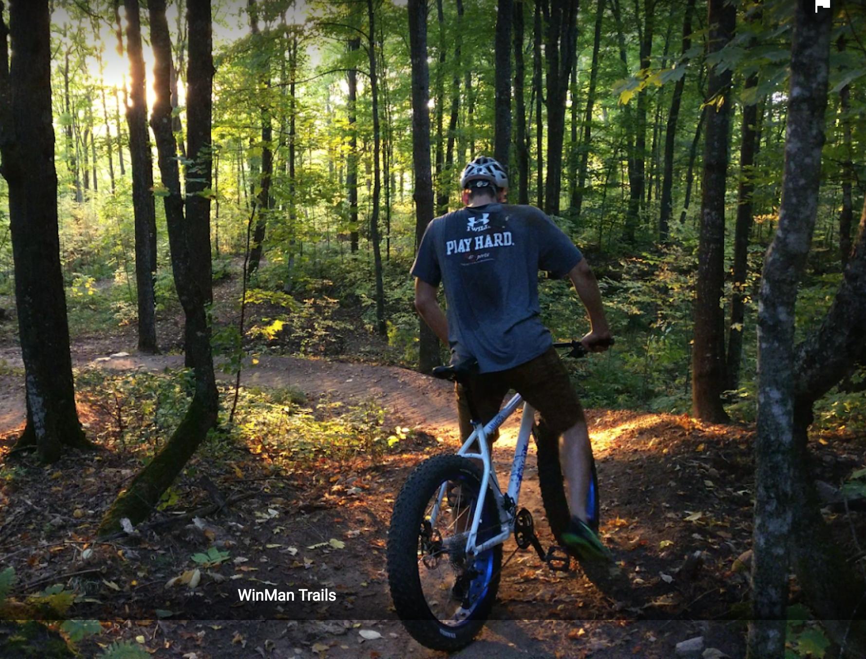 A person riding a mountain bike along a forested trail at sunset, surrounded by tall trees and lush greenery. The sunlight filters through the trees, casting a warm glow on the path. The rider is wearing a gray shirt with the words "PLAY HARD." WinMan Trails mountain bike trail.