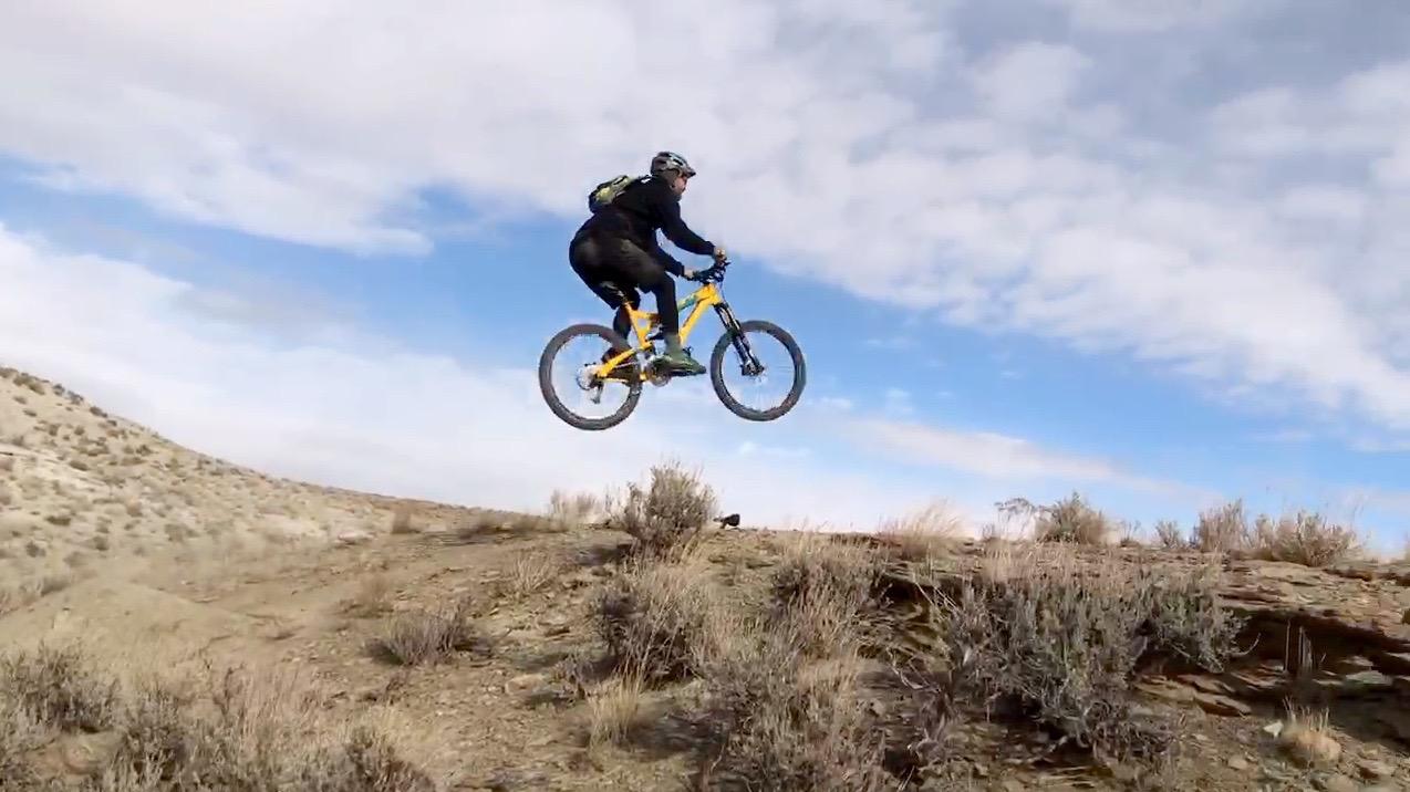 A mountain biker in mid-air making a jump over a dirt trail, surrounded by sparse vegetation against a blue sky with scattered clouds. Wilkins Peak Trails mountain bike trail.