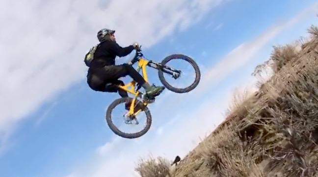 A mountain biker performing a jump over a rocky terrain with a clear blue sky in the background. The rider is wearing a helmet and black clothing, showcasing an active outdoor sports scene. Wilkins Peak Trails mountain bike trail.