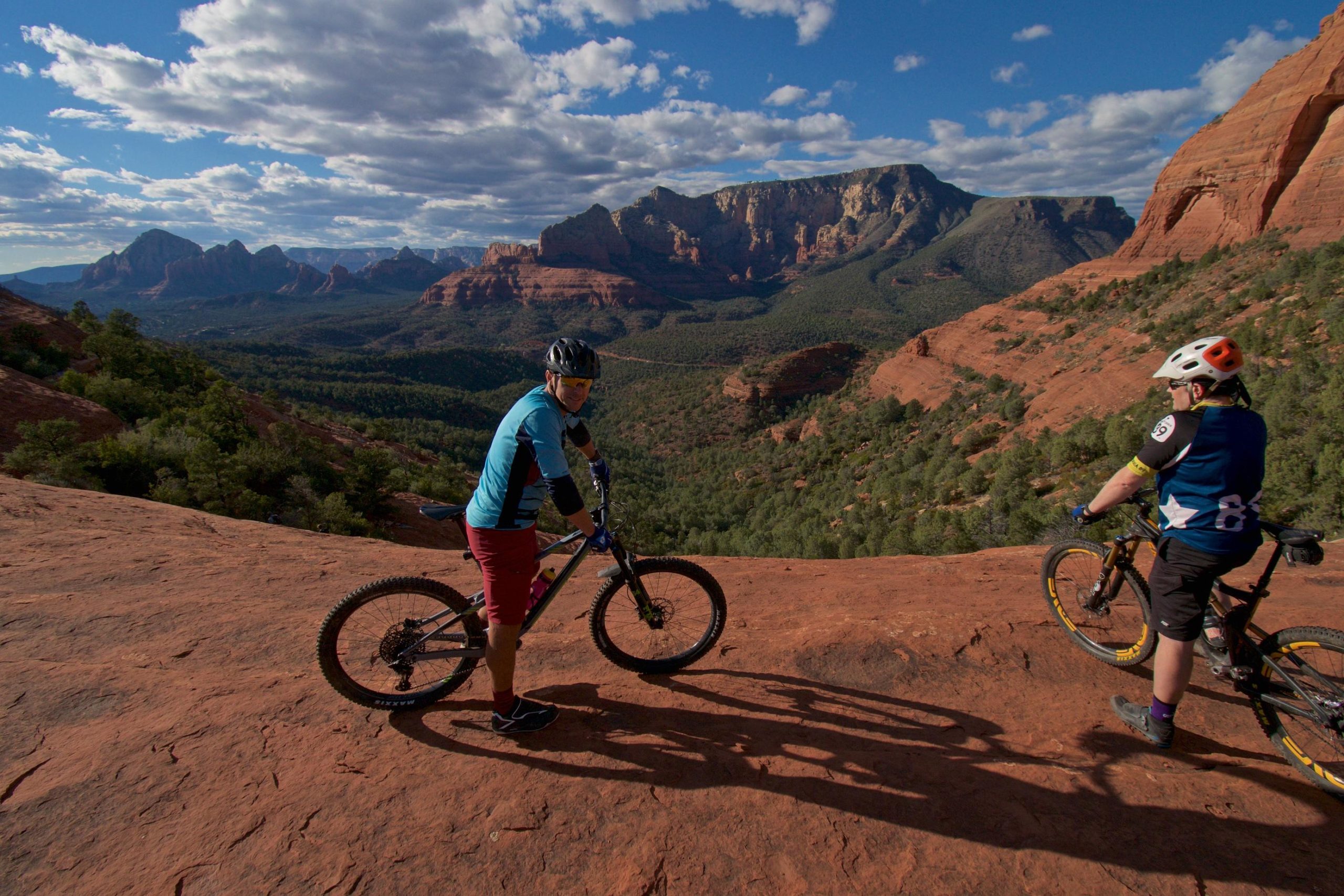 Two mountain bikers pause on a rocky overlook, surrounded by vibrant red rock formations and a lush green valley below. The sky is partly cloudy, creating a dramatic backdrop against the expansive landscape of mountains and trees. One biker stands next to their bike, while the other leans slightly forward, both enjoying the scenic view. Hangover mountain bike trail.