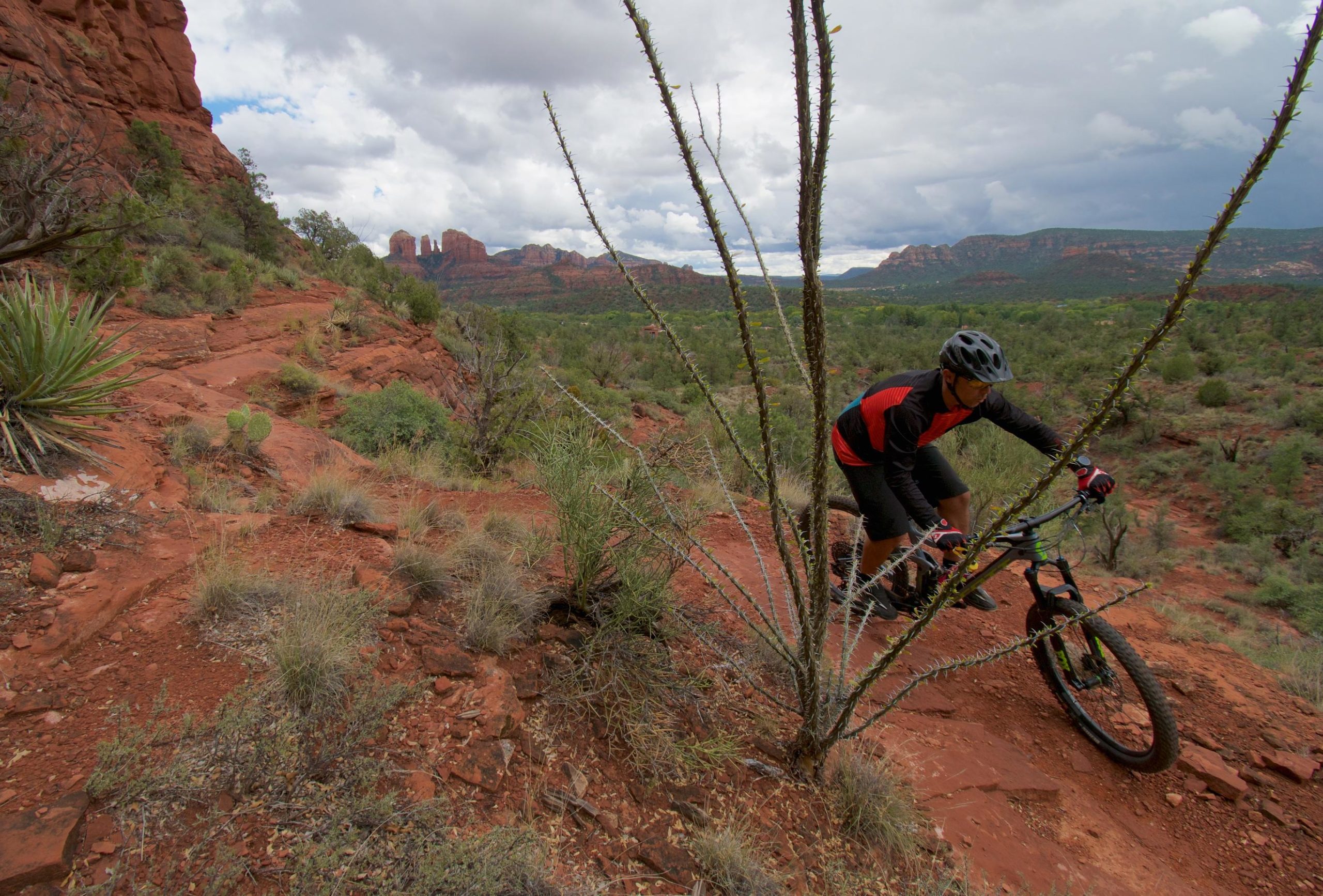 A mountain biker navigates a rocky trail in a desert landscape, surrounded by red rock formations and sparse vegetation, under a cloudy sky. Pyramid mountain bike trail.