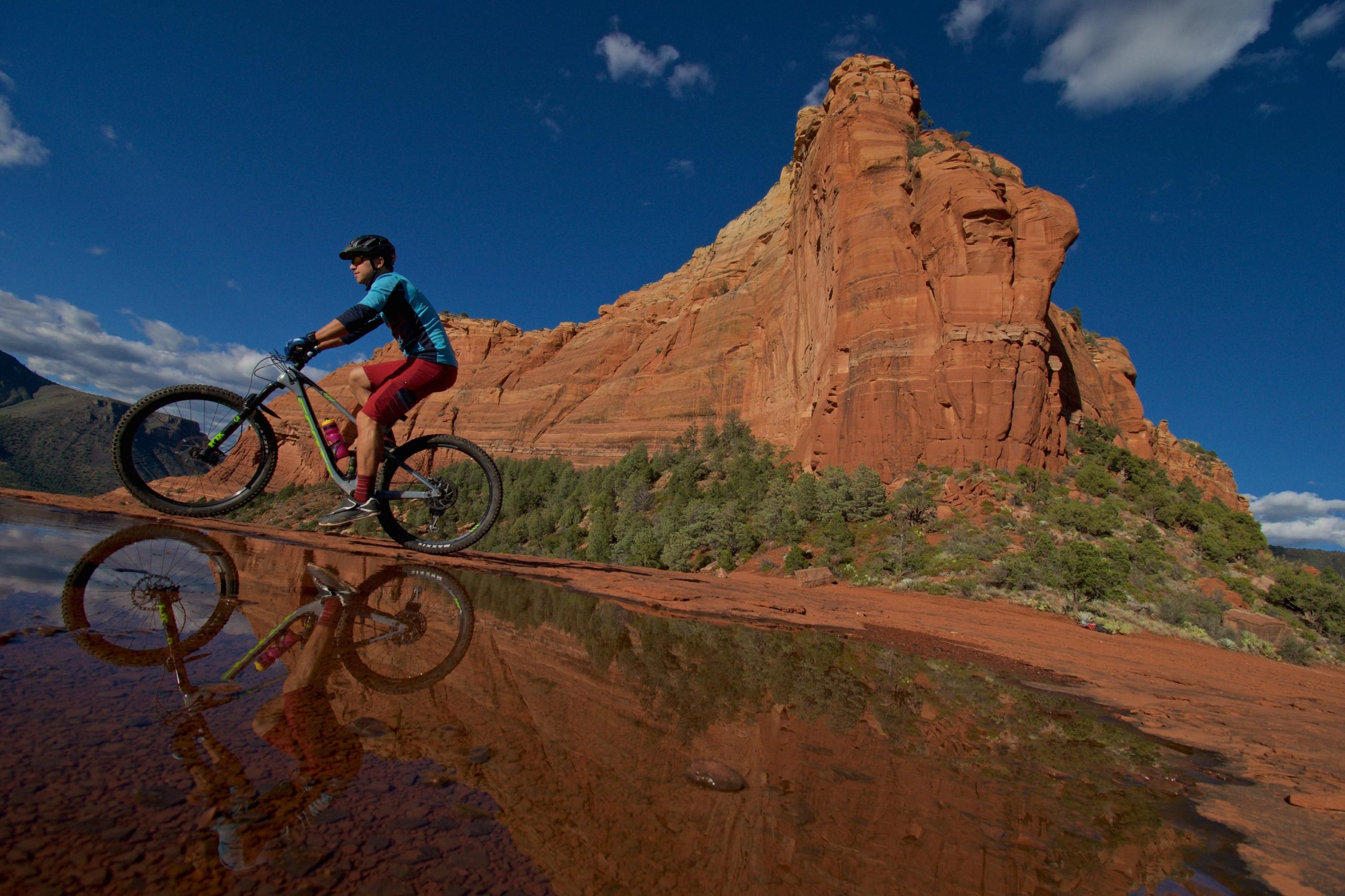 A mountain biker performing a wheelie on a rocky terrain, with vibrant red rock formations in the background and a clear blue sky overhead. The scene reflects in a shallow pool of water, showcasing the biker and the surrounding landscape. Hangover mountain bike trail.