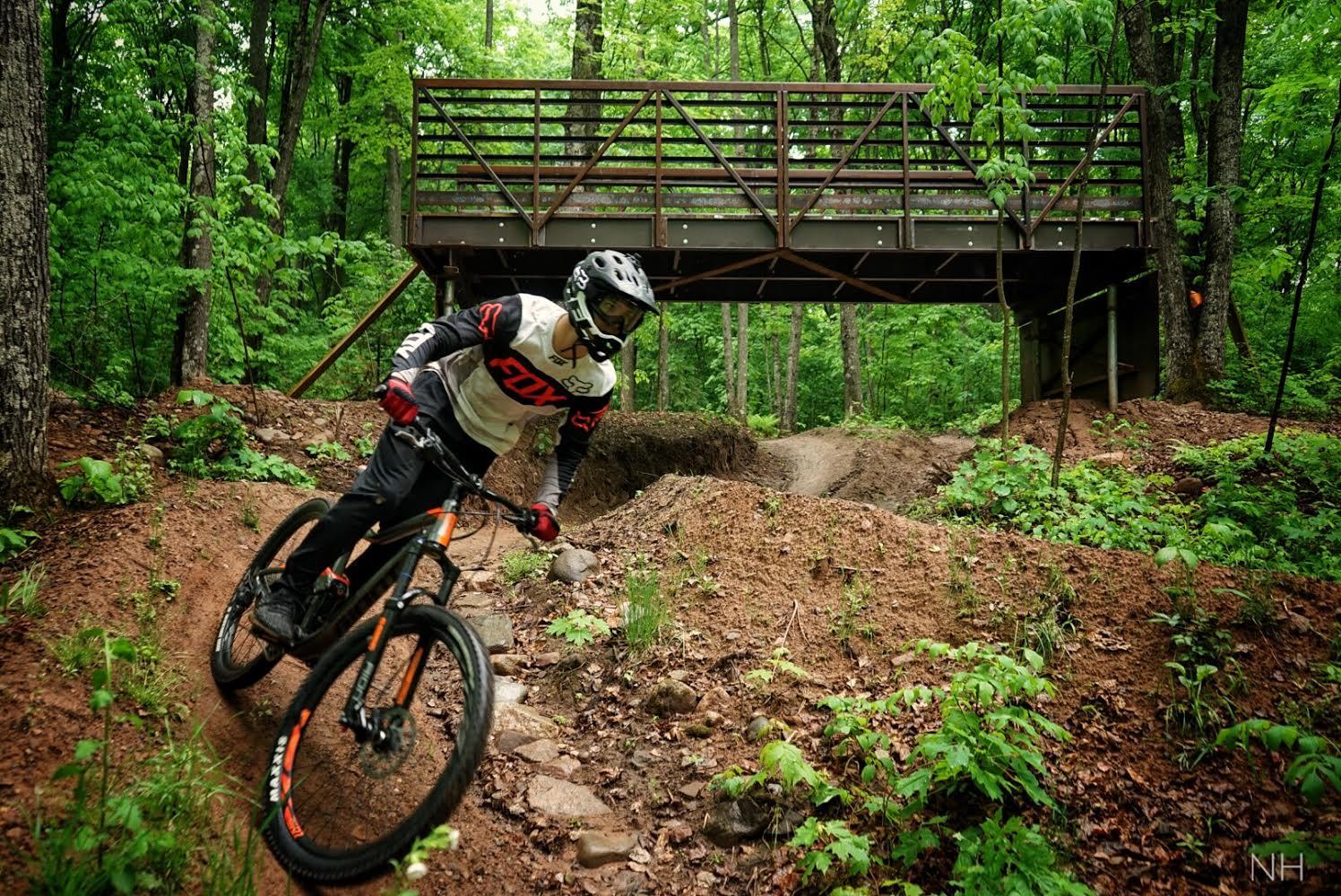 A mountain biker navigates a winding dirt trail surrounded by lush greenery, with a wooden bridge overhead in a forested area. The biker is wearing a helmet and protective gear, showcasing an action-packed moment in nature. WinMan Trail mountain bike trail.