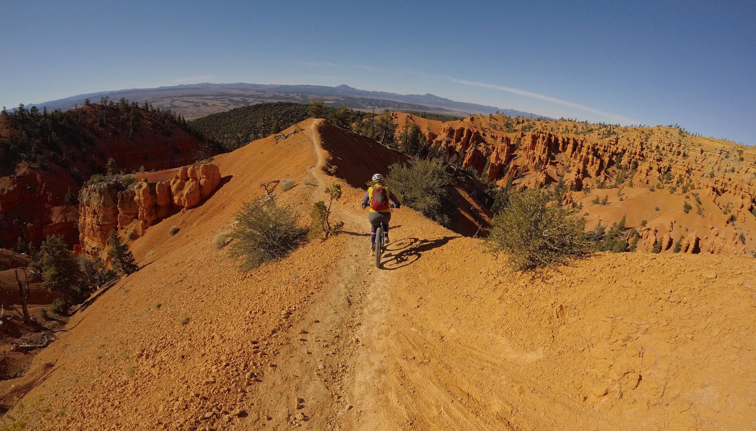 A lone mountain biker riding along a narrow trail on a bright orange ridge, surrounded by rocky cliffs and sparse trees under a clear blue sky. The landscape offers expansive views of rolling hills and distant mountains. Thunder Mountain mountain bike trail.