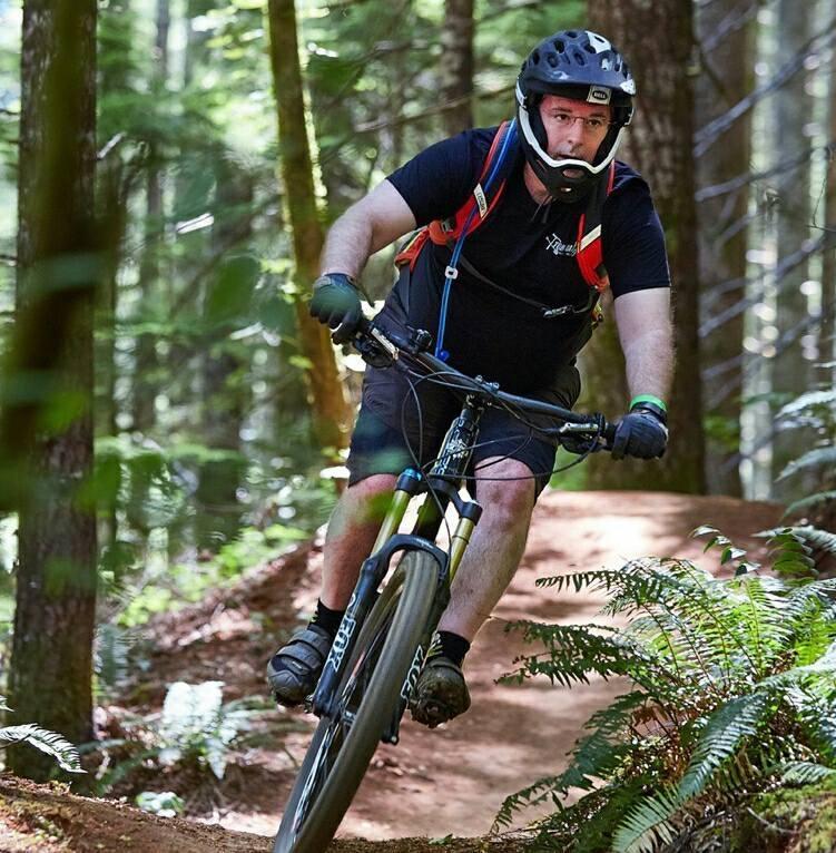 Yeti SB66: A mountain biker riding on a dirt trail through a lush forest, wearing a helmet and gloves, with a focused expression. The bike is tilted as the rider navigates a turn, surrounded by green foliage and tall trees.
