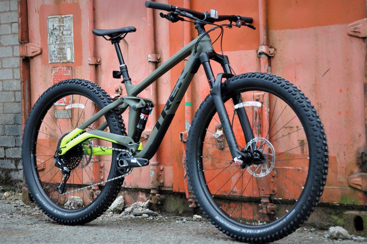 Trek Full Stache: A green mountain bike with thick, textured tires is positioned against an orange metal backdrop. The bike features a sleek design, visible suspension, and a black frame accent. The setting is urban, showcasing a mix of brick and metal surfaces.