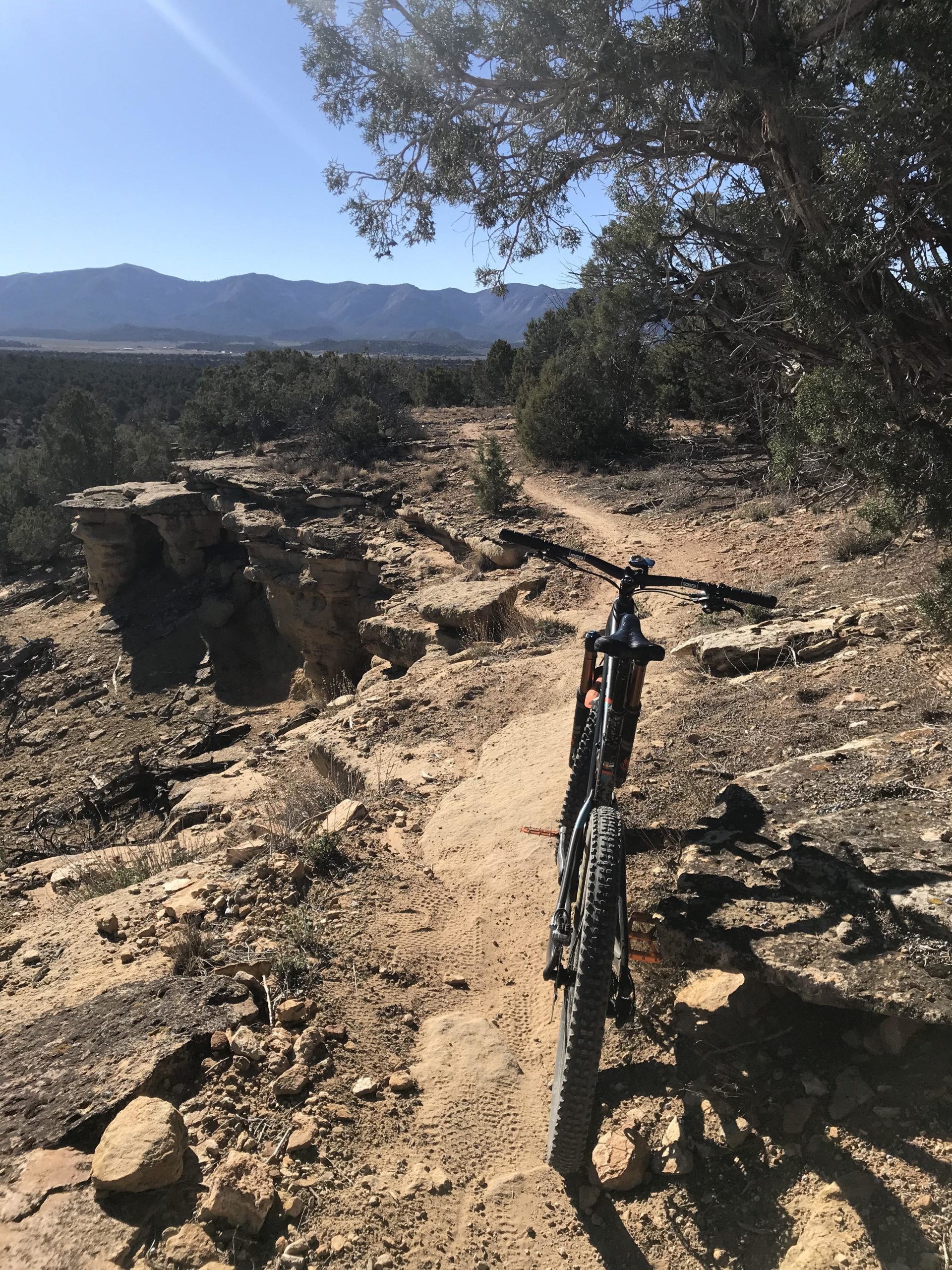 A mountain bike is positioned on a rugged trail surrounded by natural terrain, with rocky outcrops and vegetation visible. In the background, a range of mountains stretches under a clear blue sky. The path appears narrow and uneven, suggesting an adventurous biking route. Phil's World mountain bike trail.