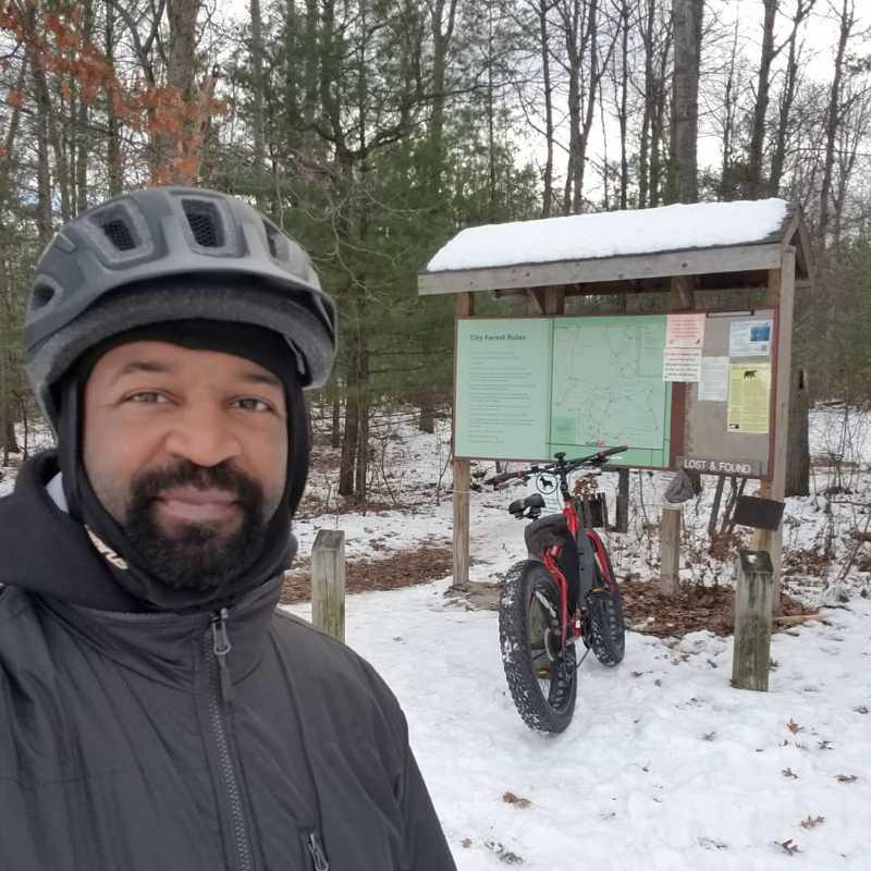 A man wearing a helmet and winter clothing smiles at the camera in a snowy forest setting. Behind him is a bike leaning against a sign featuring a map and information about city forest rules. Snow covers the ground, and trees are visible in the background. Midland City Forest mountain bike trail.