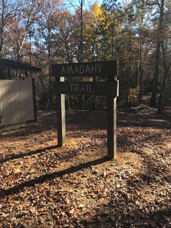 Wooden trail sign reading "Amadahy Trail" at the entrance, surrounded by autumn foliage and trees. A gravel path can be seen leading into the wooded area. Woodring Branch mountain bike trail.