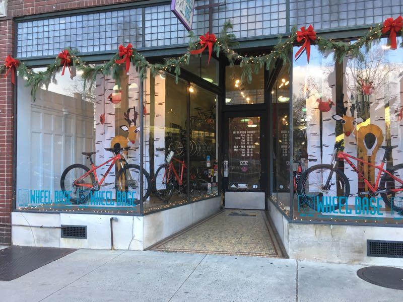 Storefront of a bicycle shop decorated for the holidays, featuring two red bicycles in the window with a festive display of holiday decor, including wreaths and ornaments. The shop name "WHEEL BASE" is visible on the window, and the entrance is adorned with a welcoming ambiance.