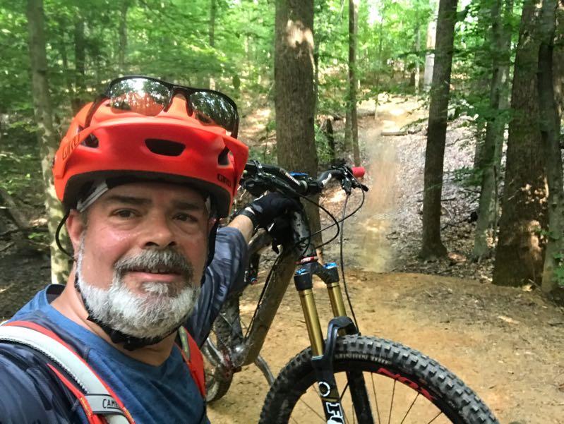 A man in a red helmet and sunglasses smiles for a selfie while standing next to his mountain bike on a forest trail. The background features lush green trees and a dirt path, indicating a biking or hiking environment. Fountainhead Regional Park mountain bike trail.