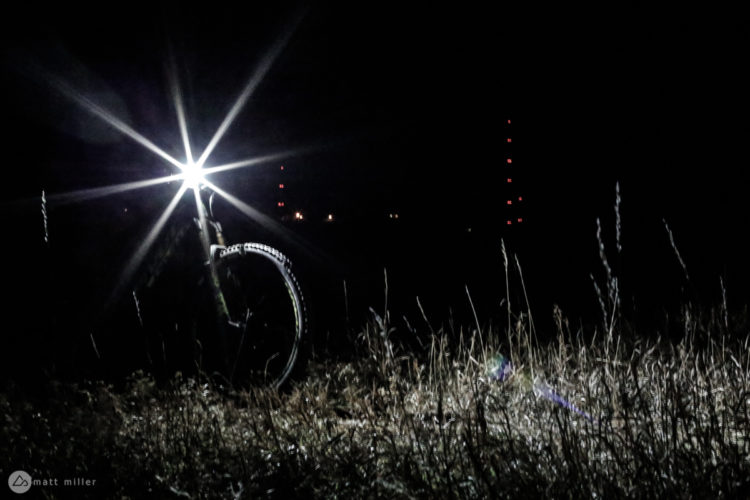 A mountain bike with a bright front light stands in tall grass at night, illuminating the surrounding area. In the background, faint red lights of communication towers are visible against the dark sky.